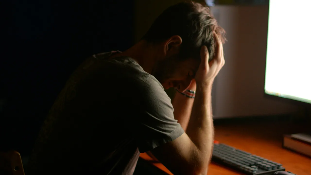 Bărbat sub presiune, cu mâinile la cap, în fața unui monitor aprins într-un birou întunecat
